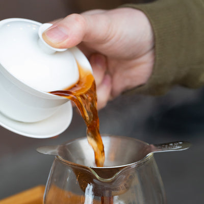 Hand pouring brewed tea out of gaiwan vessel through strainer into tea decanter.