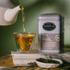 A silver tin of Smith Teamaker No. 17 2nd Flush Darjeeling tea stands beside a glass cup with tea being poured from a white teapot, with loose tea leaves and a small dish in the foreground against a muted green wall.