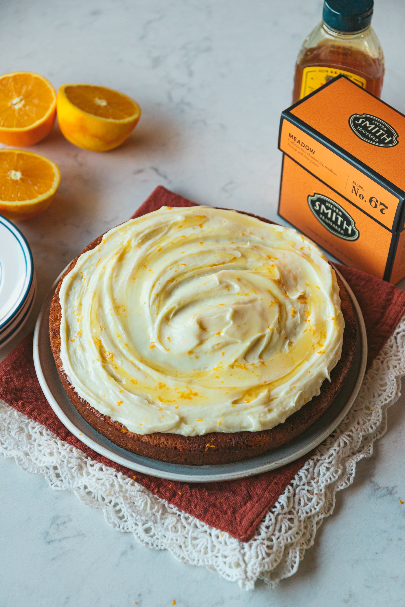 Cake with swirls of white frosting, honey and orange zest on top next to a box of Meadow herbal infusion on a countertop. 