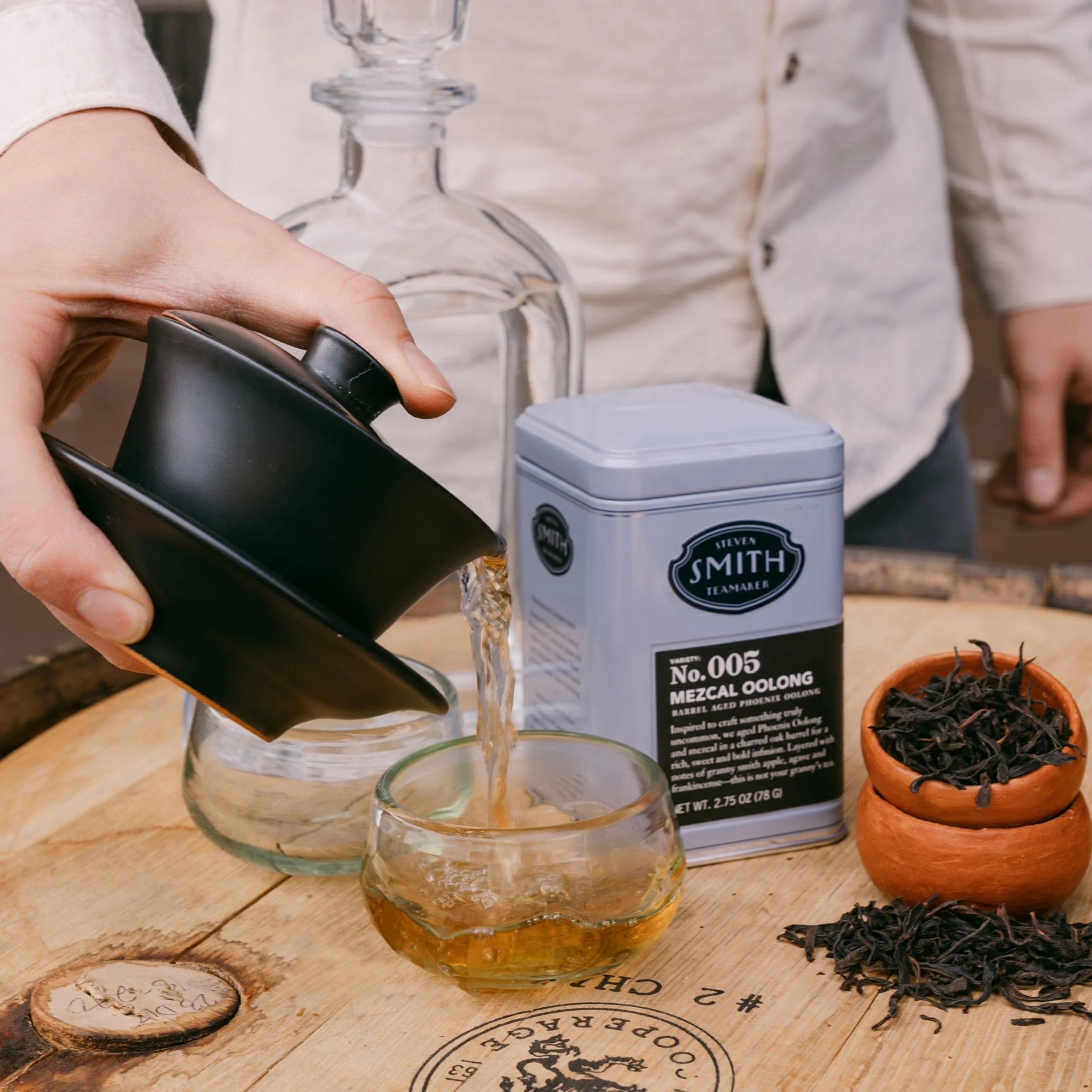 Person pouring tea into a glass with a Smith tea canister and tea leaves on a wooden surface.