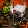 Mezcal Oolong tea canister labeled 'Smith' with a glass of tea and potted plants on a wooden surface