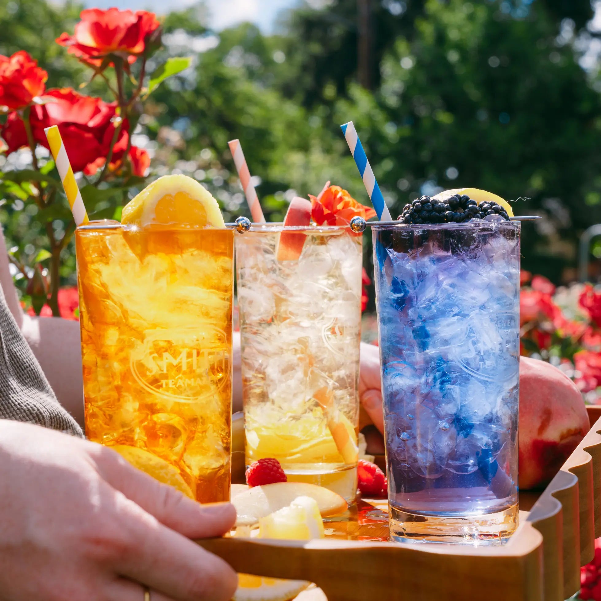 Three tall glasses of colorful iced drinks on a wooden tray outdoors, with a hand holding the tray. The drinks are amber, clear with lemon and raspberries, and blue-purple with blackberries, each with a striped straw.