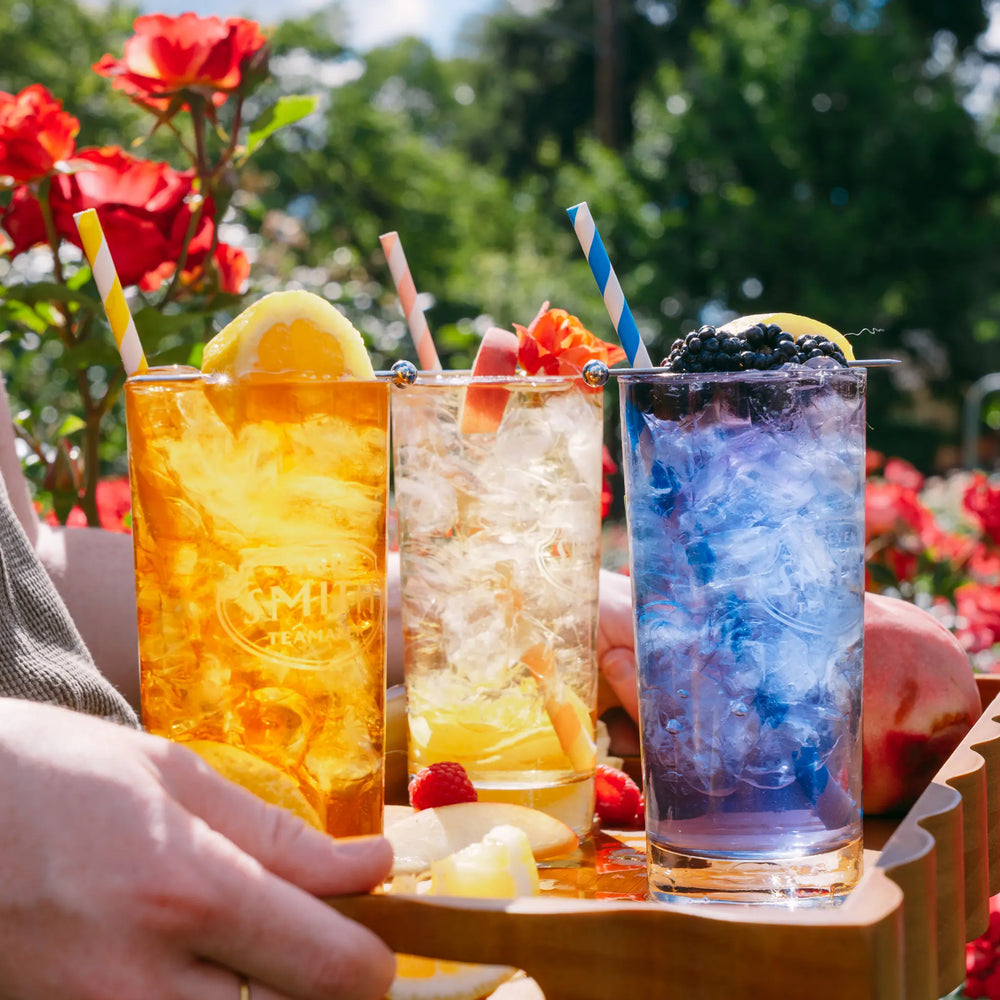 Three tall glasses of colorful iced drinks on a wooden tray outdoors, with a hand holding the tray. The drinks are amber, clear with lemon and raspberries, and blue-purple with blackberries, each with a striped straw.