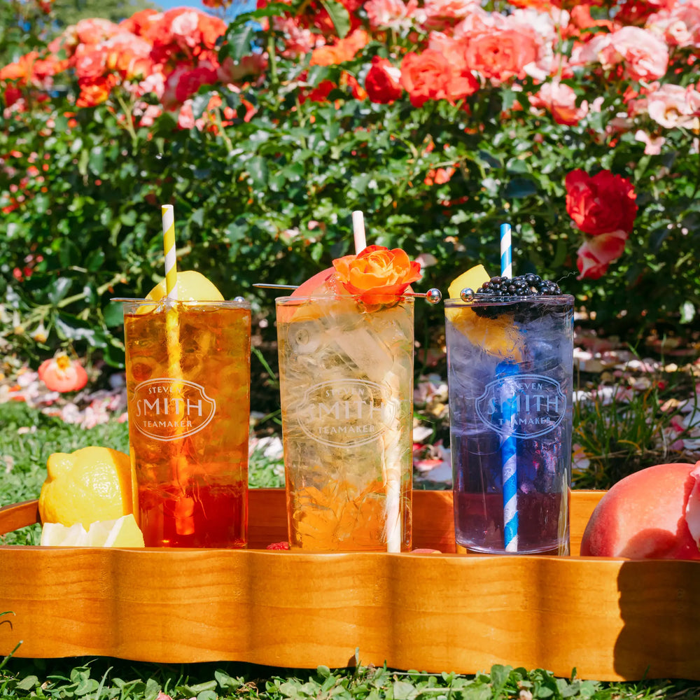 Three tall clear cups filled with colorful iced drinks, arranged in a wooden tray outdoors on grass with flowers and greenery in the background. The cups have visible 'SMITH COLLECTION' branding.