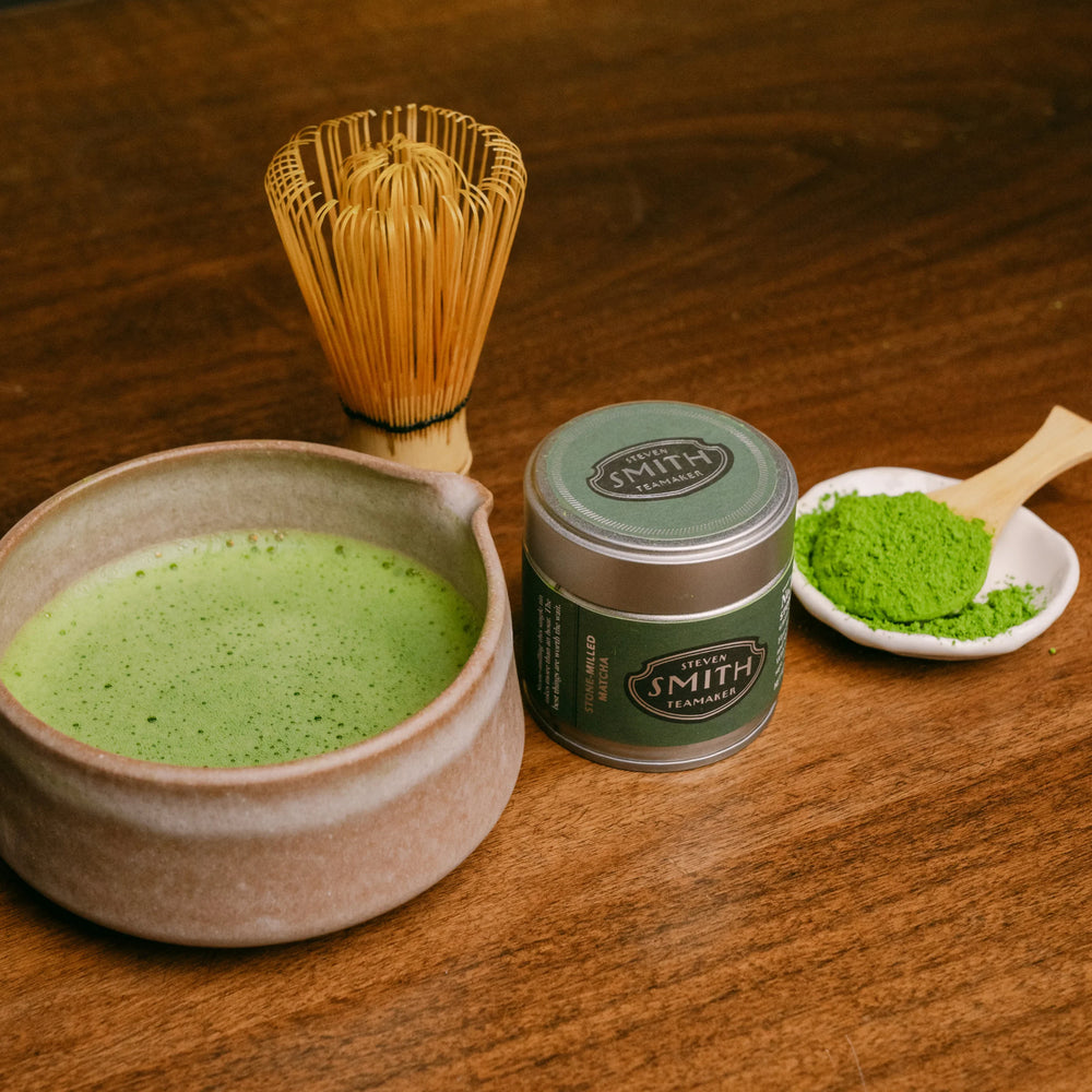 Tea-making setup with a bowl of matcha tea, matcha whisk, and container of Smith Tea on a wooden surface.
