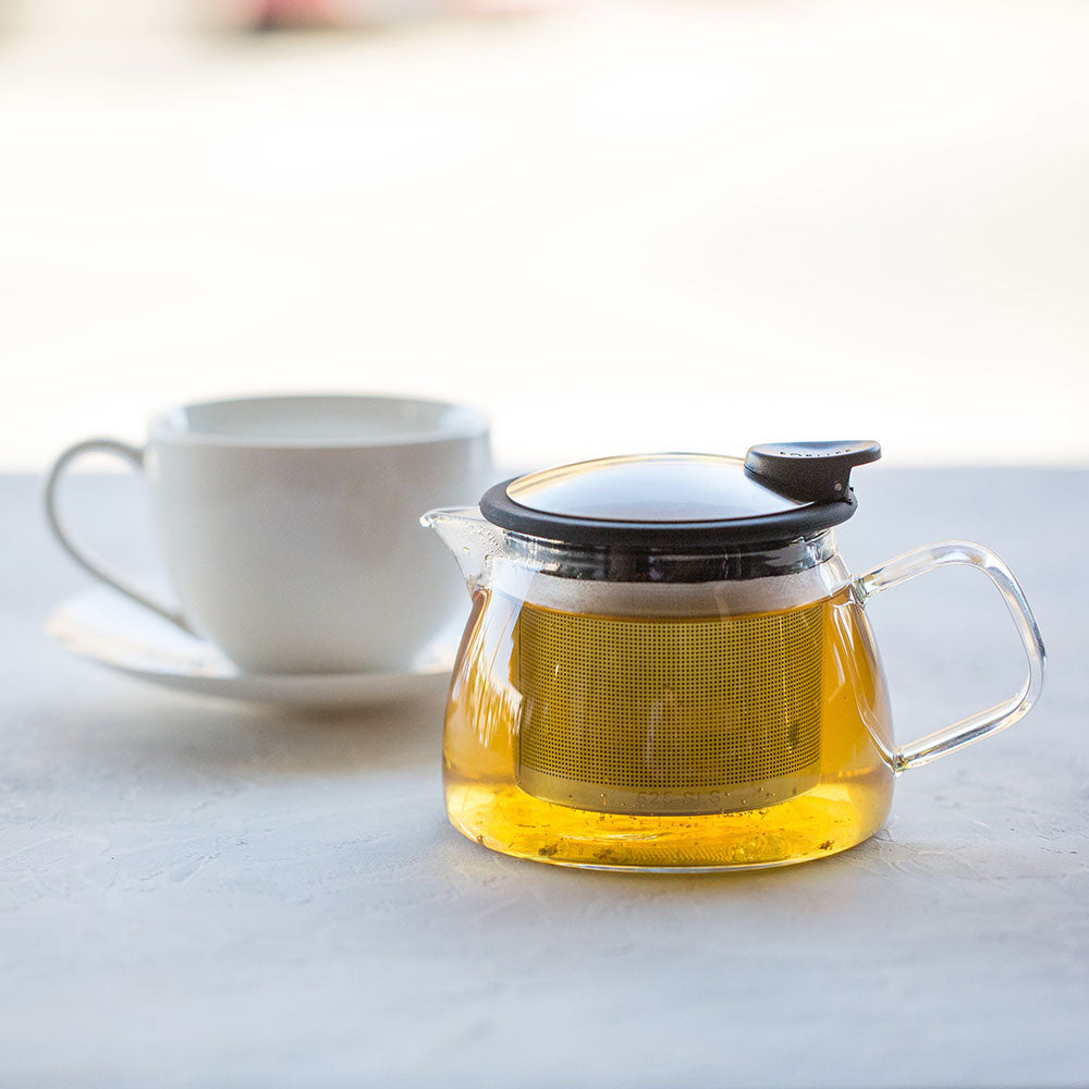 Glass teapot with stainless steel and black lid with spout and handle filled with amber colored tea.