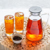 Person pouring glass pitcher with black lid filled with light brown iced tea.