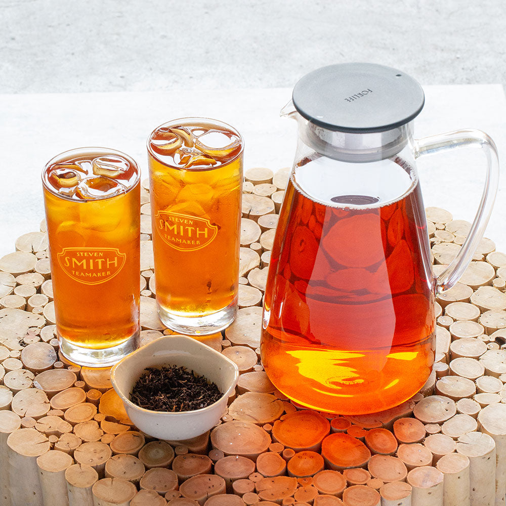 Person pouring glass pitcher with black lid filled with light brown iced tea.