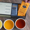 Dark silver box with black trim slightly opened to display orange and black tea sachets inside.